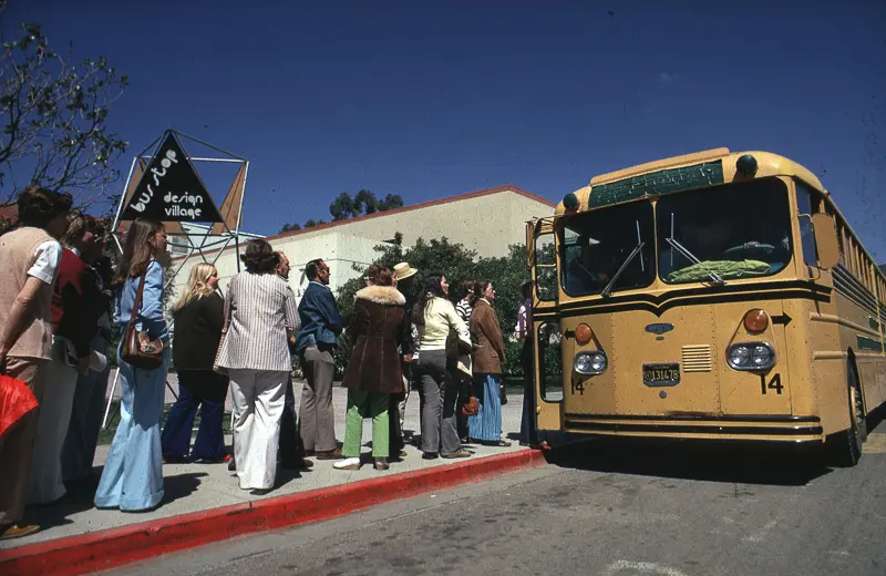 Students gathering beside a yellow bus and a Design Village sign.