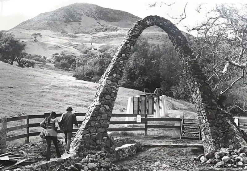 Two people walking through the entry arch toward the stone wall, old information signs present.