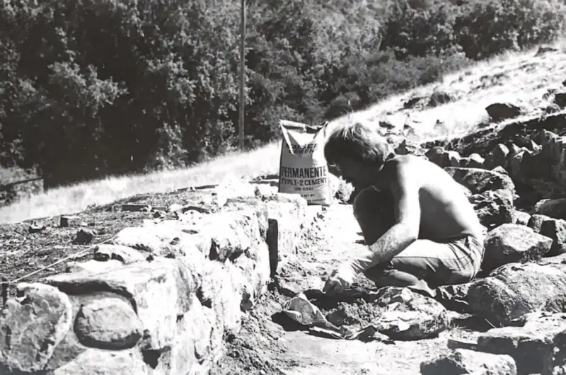 A builder constructing the stone wall extending from the arch.