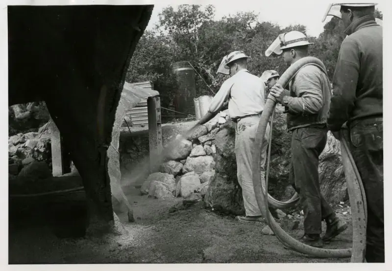A worker spraying concrete on one of the three contact points while others observe.